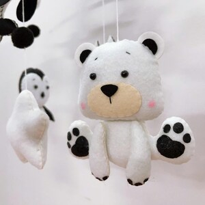 K&ouml;nnte beinhalten: Handgefertigtes Babymobile mit Filztierfiguren. Das Mobile enth&auml;lt einen wei&szlig;en B&auml;ren mit schwarzen Pfotenabdr&uuml;cken, einen Panda und einen Pinguin. Der B&auml;r hat eine hellbraune Schnauze und rosa Wangen. Die Figuren sind an wei&szlig;en Schn&uuml;ren aufgeh&auml;ngt.