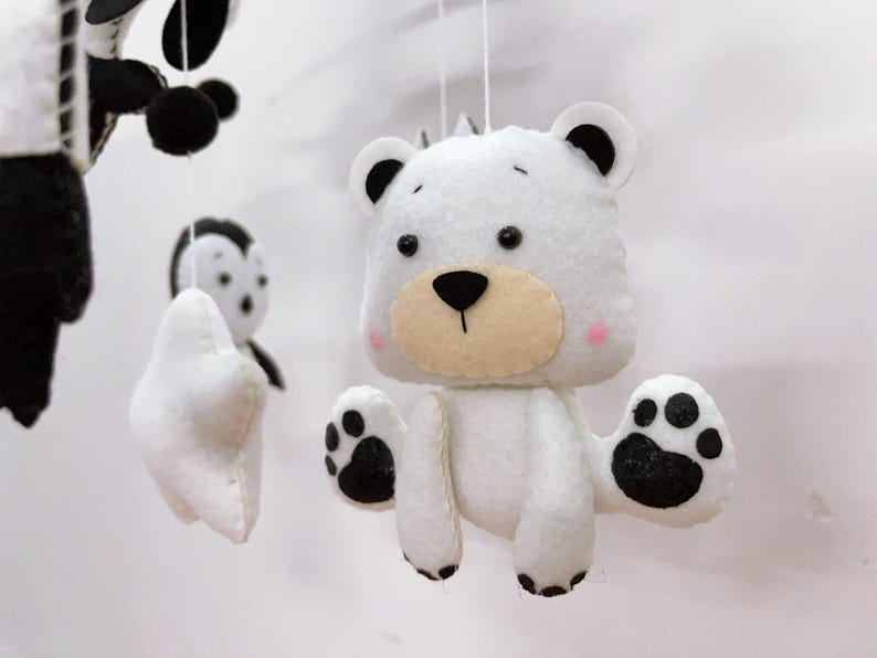K&ouml;nnte beinhalten: Handgefertigtes Babymobile mit Filztierfiguren. Das Mobile enth&auml;lt einen wei&szlig;en B&auml;ren mit schwarzen Pfotenabdr&uuml;cken, einen Panda und einen Pinguin. Der B&auml;r hat eine hellbraune Schnauze und rosa Wangen. Die Figuren sind an wei&szlig;en Schn&uuml;ren aufgeh&auml;ngt.