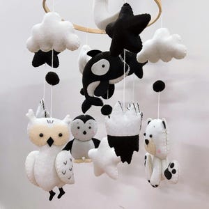 K&ouml;nnte beinhalten: Handgefertigtes Baby-Mobile mit Filztieren in Schwarz und Wei&szlig;. Das Mobile enth&auml;lt eine Eule, einen Pinguin, einen Eisb&auml;ren, einen Wal, Wolken, Sterne und eine Krone, die an einem Holzreifen aufgeh&auml;ngt sind.