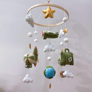 Puede incluir: Un m&oacute;vil colgante para habitaci&oacute;n infantil con un marco circular de madera y adornos de fieltro que incluyen una estrella dorada, un avi&oacute;n verde, un cohete, un globo aerost&aacute;tico, un globo terr&aacute;queo, una maleta con un icono de avi&oacute;n y nubes blancas.