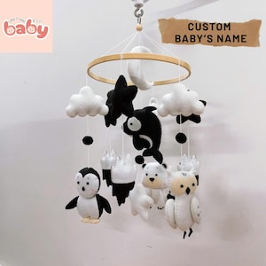 K&ouml;nnte beinhalten: Ein Baby-Mobile mit Tierfiguren aus Filz in Schwarz, Wei&szlig; und Creme. Das Mobile enth&auml;lt einen Pinguin, eine Eule, einen Eisb&auml;ren, einen Wal, Wolken, Sterne und einen Mond, die an einem Holzreifen aufgeh&auml;ngt sind. Der Text "CUSTOM BABY'S NAME" ist sichtbar.