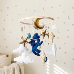 Puede incluir: Un móvil colgante con un dragón central de fieltro azul oscuro rodeado de lunas crecientes doradas metálicas, estrellas doradas y blancas, nubes de fieltro blanco, un huevo moteado color cerceta y antorchas de fieltro beige.