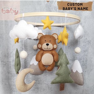 Puede incluir: Móvil para bebé con un oso pardo, estrellas amarillas, nubes blancas y árboles verdes. El móvil está suspendido de un aro de madera y lleva el texto "CUSTOM BABY'S NAME". Ideal para una habitación infantil.