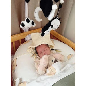 K&ouml;nnte beinhalten: Ein schlafendes Baby in einem beigen Hut und Pyjama, eingebettet in ein wei&szlig;es und beige gemustertes Babybett. &Uuml;ber dem Baby h&auml;ngt ein Mobile mit schwarzen und wei&szlig;en Filzformen. Das Bett befindet sich in einem Holzbettchen.