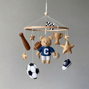 Puede incluir: Móvil de bebé hecho a mano con un oso de peluche con una camiseta azul con una "C" blanca, rodeado de adornos de fieltro con temática deportiva. Incluye un bate de béisbol, fútbol americano, balón de fútbol, trofeo, estrellas y una zapatilla en miniatura. Cuelga de un aro de madera.