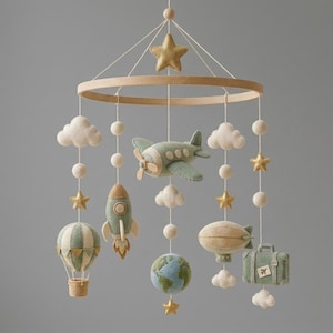 Puede incluir: Un m&oacute;vil colgante para habitaci&oacute;n infantil con figuras de fieltro suspendidas de un marco circular de madera. Incluye un avi&oacute;n verde salvia, un cohete, un globo aerost&aacute;tico, un globo terr&aacute;queo, una maleta, nubes y estrellas doradas.