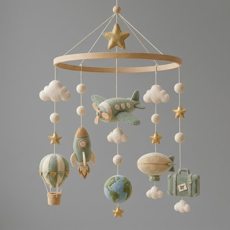 Puede incluir: Un m&oacute;vil colgante para habitaci&oacute;n infantil con figuras de fieltro suspendidas de un marco circular de madera. Incluye un avi&oacute;n verde salvia, un cohete, un globo aerost&aacute;tico, un globo terr&aacute;queo, una maleta, nubes y estrellas doradas.