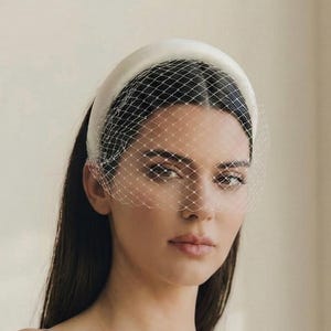 May include: A thick, smooth, off-white padded headband featuring an attached delicate white birdcage veil with a diamond-shaped netting pattern that covers the upper portion of the face, creating a structured and layered headpiece accessory.