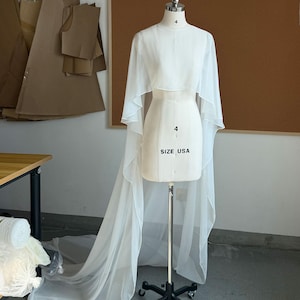 May include: A white dress form with a sheer, flowing cape and train. The dress form is labeled "SIZE USA 4". The cape has a high neckline and extends into long, elegant panels. The dress form is on a black wheeled stand.