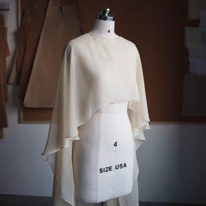 May include: A cream-colored, sheer, flowing cape-style garment draped over a white dress form. The form features the text 4 and SIZE USA printed on the front. The cape has a high-low hemline with soft, layered fabric panels that extend down.