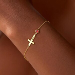 May include: A gold-colored bracelet featuring a cross pendant with the name "Clare" engraved on it. The bracelet also includes a small, round, red gemstone. The delicate chain is attached to the cross and gemstone.
