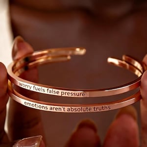 Puede incluir: Brazaletes de puño de color oro rosa. El brazalete superior dice "worry fuels false pressure" y el inferior "emotions aren't absolute truths". Los brazaletes son lisos y minimalistas.