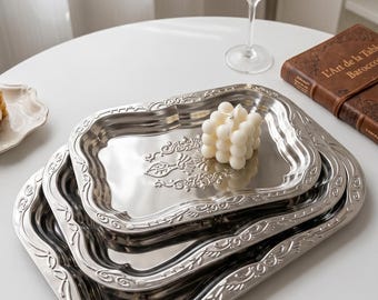 May include: Set of three nested silver trays with ornate, embossed floral and scrollwork designs. A white bubble candle sits on the top tray. A glass of champagne and a book are in the background.
