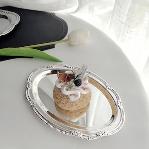 May include: A silver oval tray with ornate edges holds a small, round dessert. The dessert is topped with white frosting, dried fruit, and berries. A small tag is attached. A white tulip and black placemat are in the background.