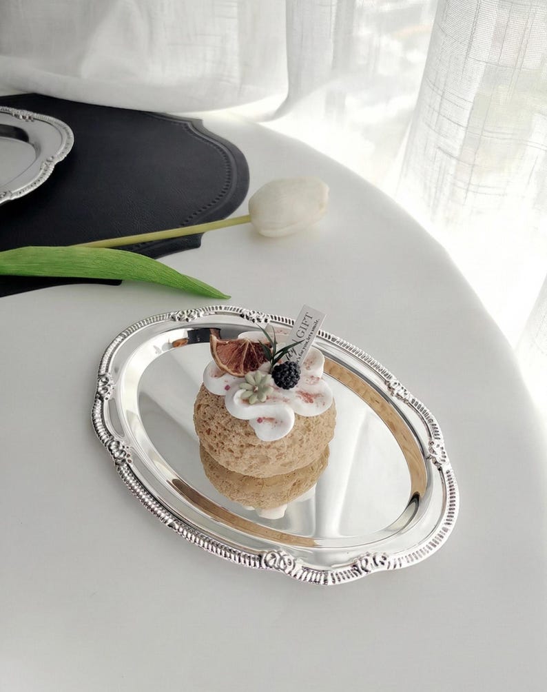 May include: A silver oval tray with ornate edges holds a small, round dessert. The dessert is topped with white frosting, dried fruit, and berries. A small tag is attached. A white tulip and black placemat are in the background.