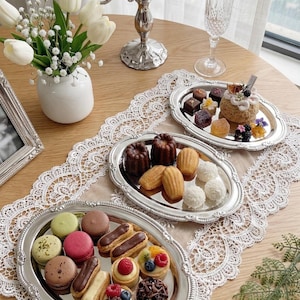 May include: A table setting with various desserts on silver platters. The platters hold macarons, eclairs, and mini tarts. A silver candelabra and a vase of white tulips are also present, creating an elegant display.