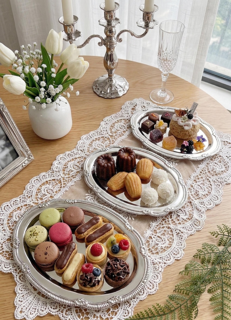 May include: A table setting with various desserts on silver platters. The platters hold macarons, eclairs, and mini tarts. A silver candelabra and a vase of white tulips are also present, creating an elegant display.