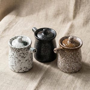 May include: Three ceramic condiment jars with lids. The jars are speckled in white, black, and brown. Each jar has a small handle and a lid with a knob. The jars are arranged on a neutral-coloured fabric.