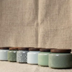 Peut inclure: Une collection de pots en céramique avec des couvercles en bois. Les pots sont de différentes couleurs, dont le blanc, le vert et un motif craquelé. Les pots sont disposés en rangée, mettant en valeur leurs différentes textures et finitions.