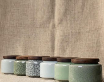 May include: A collection of ceramic jars with wooden lids. The jars are in various colors, including white, green, and a crackle glaze design. The jars are arranged in a row, showcasing their different textures and finishes.