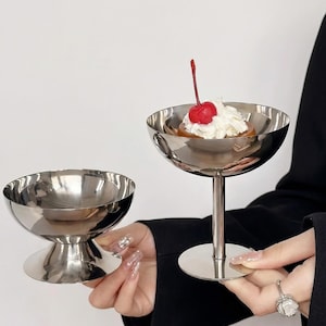 Peut inclure: Deux coupes à dessert argentées. L'une est vide, l'autre contient un dessert garni de crème fouettée et d'une cerise rouge. Les coupes ont un socle et une forme arrondie. La coupe à dessert est tenue par une personne.