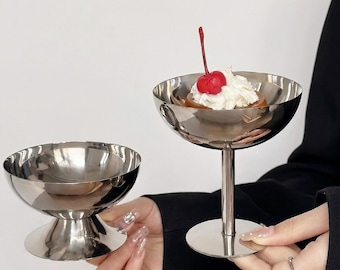 May include: Two silver-colored dessert bowls. One bowl is empty, and the other contains a dessert topped with whipped cream and a red cherry. The bowls have a pedestal base and a rounded bowl shape. The dessert bowl is held by a person.