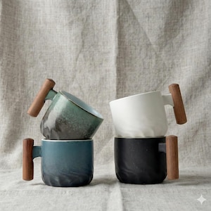 Peut inclure: Quatre tasses en céramique avec des poignées en bois sont empilées. Les tasses sont dans des tons de bleu sarcelle, blanc, bleu et noir. Les poignées sont en bois. Les tasses sont sur un fond en tissu de couleur neutre.