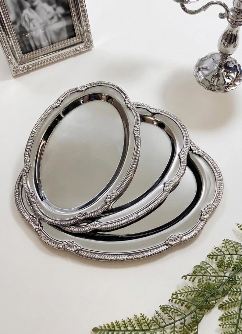 May include: Three oval, silver-colored serving trays with ornate, scalloped edges. The trays are stacked, with the top one in focus. A silver picture frame and candlestick holder are in the background.