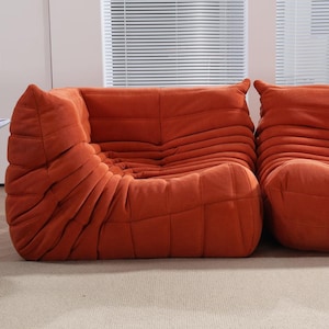 Puede incluir: Dos sof&aacute;s modulares de color naranja quemado con un dise&ntilde;o acolchado texturizado. Los sof&aacute;s tienen un perfil bajo y bordes redondeados, con una apariencia suave y acogedora. El fondo incluye una ventana con persianas y una alfombra de color neutro.