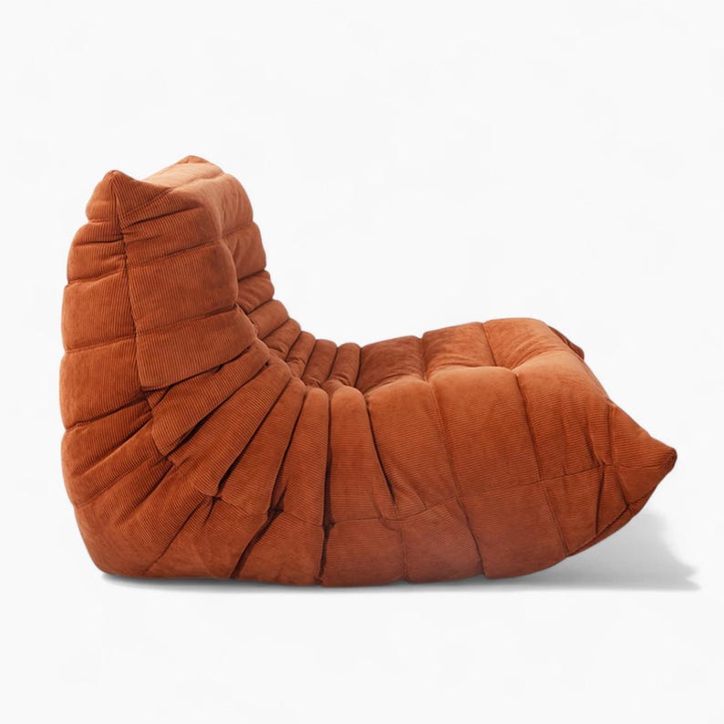 Puede incluir: Un sill&oacute;n lounge de pana color &oacute;xido con un dise&ntilde;o curvo &uacute;nico. La silla presenta una serie de pliegues verticales a lo largo del respaldo y el asiento, creando una apariencia texturizada. La silla est&aacute; dise&ntilde;ada para la comodidad y la relajaci&oacute;n.