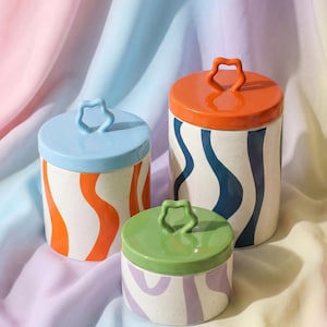 Puede incluir: Tres botes de cerámica con tapas. Los botes son blancos con rayas onduladas de colores en naranja, azul y morado. Cada tapa tiene un asa en forma de estrella y está coloreada en naranja, azul y verde.