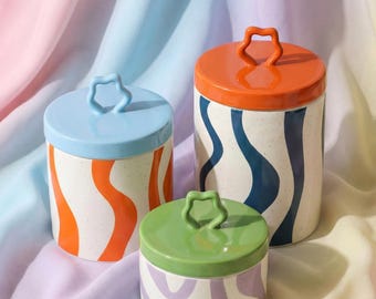 May include: Three ceramic canisters with lids. The canisters are white with colorful wavy stripes in orange, blue, and purple. Each lid has a star-shaped handle and is colored in orange, blue, and green.