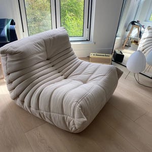 Puede incluir: Un sill&oacute;n lounge de pana beige con un perfil bajo y una forma redondeada. La silla tiene una serie de crestas horizontales y un aspecto suave y acogedor. La silla est&aacute; en una habitaci&oacute;n bien iluminada con una ventana y un espejo.