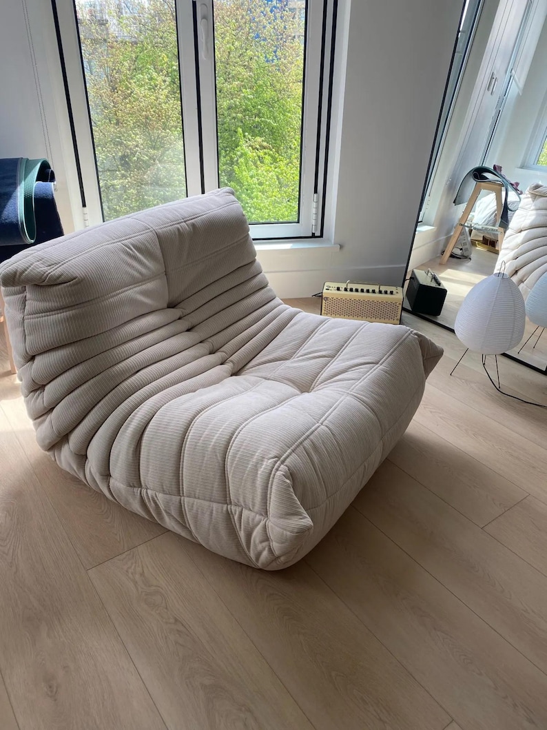 Puede incluir: Un sill&oacute;n lounge de pana beige con un perfil bajo y una forma redondeada. La silla tiene una serie de crestas horizontales y un aspecto suave y acogedor. La silla est&aacute; en una habitaci&oacute;n bien iluminada con una ventana y un espejo.