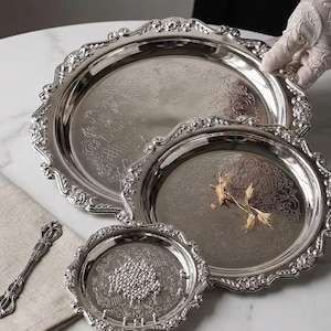 May include: A collection of three silver-plated serving trays with decorative floral borders and intricate engraved designs. The trays are of varying sizes, with a small decorative spoon on a linen napkin. A gloved hand is holding one of the trays.