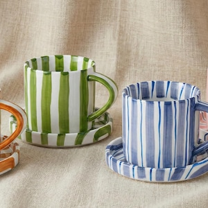 Peut inclure: Quatre tasses en céramique avec soucoupes assorties, chacune avec un motif à rayures verticales. Les tasses sont orange, vertes, bleues et roses. Les tasses ont des anses et sont posées sur une surface neutre.
