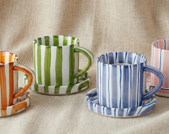 May include: Four ceramic mugs with matching saucers. Each mug features a vertical striped pattern in orange, green, blue, and pink. The mugs have rounded handles and are set on a neutral-colored surface.