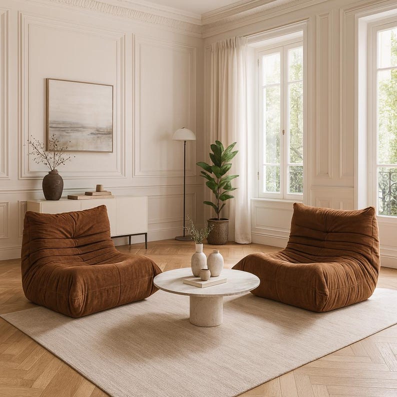 Puede incluir: Un sal&oacute;n acogedor con dos sillones bajos de pana marr&oacute;n. Una mesa de centro redonda y blanca se encuentra entre las sillas sobre una alfombra de tonos neutros. La habitaci&oacute;n cuenta con grandes ventanales, una l&aacute;mpara de pie y jarrones decorativos.