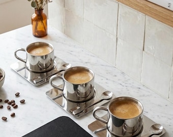 May include: Three stainless steel espresso cups filled with coffee, each on a matching square saucer with a spoon. The cups sit on a white marble countertop, with coffee beans scattered nearby. A black placemat is in the foreground.