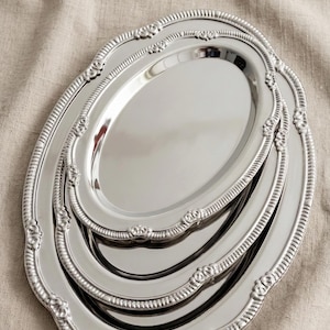 May include: A set of three oval, silver-colored serving trays. Each tray has a decorative, scalloped edge with floral accents. The trays are nested together, showcasing their varying sizes and reflective surfaces.