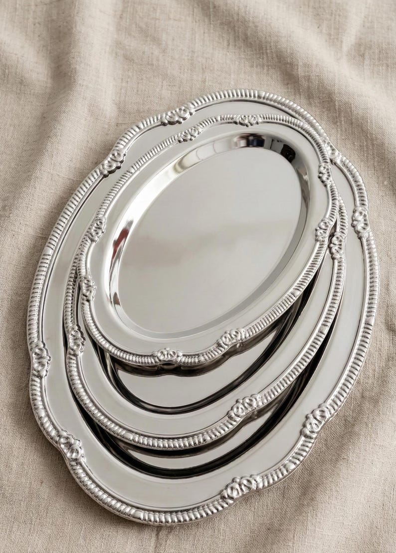 May include: A set of three oval, silver-colored serving trays. Each tray has a decorative, scalloped edge with floral accents. The trays are nested together, showcasing their varying sizes and reflective surfaces.