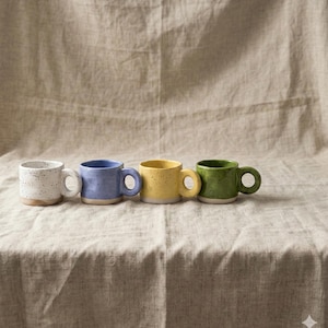 Peut inclure: Quatre petits mugs en céramique alignés, chacun avec une couleur et une finition mouchetée différentes. Les couleurs sont blanc, bleu, jaune et vert. Chaque mug a une anse arrondie et une base de couleur claire.