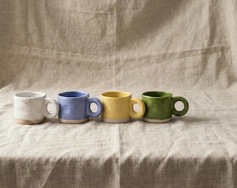 May include: Four small ceramic mugs in a row, each with a different color and speckled finish. The colors are white, blue, yellow, and green. Each mug has a rounded handle and a light-colored base.