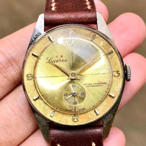 Op de afbeelding: Vintage Lucerne polshorloge met een goudkleurige wijzerplaat en een bruine leren band. De wijzerplaat toont de merknaam, een kleine secondenwijzer en de tekst "Lifetime Mainspring, Antimagnetic". Het horloge heeft zilverkleurige wijzers en uurmarkeringen.