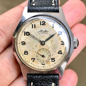 Op de afbeelding: Vintage Mido Multifort polshorloge met een zilverkleurige kast en een zwarte leren band. De wijzerplaat is gebroken wit met zwarte cijfers en wijzers. Een kleine secondenwijzer staat onderaan. Het horloge heeft een getextureerde, verouderde uitstraling.