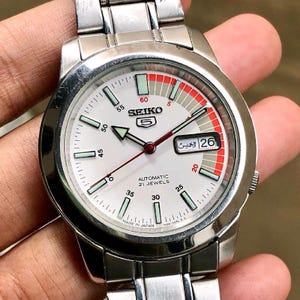 May include: A silver Seiko 5 automatic wristwatch with a white face, featuring black numerals, green and red accents, and a day-date display. The watch has a stainless steel band and is marked "Made in Japan."
