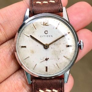 May include: Vintage Citizen wristwatch with a white face, silver case, and brown leather strap. The watch face features gold-tone hands and markers, with the brand name "CITIZEN" and the number 12 visible. The watch shows signs of age.
