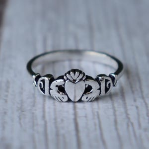 Op de afbeelding: Een zilverkleurige Claddagh-ring. De ring heeft een hart dat door twee handen wordt vastgehouden, bekroond met een kroon. De ring staat tegen een lichtgekleurde, getextureerde achtergrond.