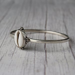 Puede incluir: Un brazalete de plata con una concha blanca como pieza central. La concha está engastada en una carcasa decorativa de plata. El brazalete tiene un diseño sencillo y elegante, adecuado para el uso diario.