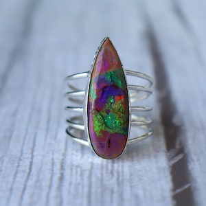 May include: A silver ring featuring a large, teardrop-shaped gemstone with a vibrant display of colors. The gemstone showcases a mix of purple, green, and red hues. The ring has a multi-band design, with several parallel silver bands.